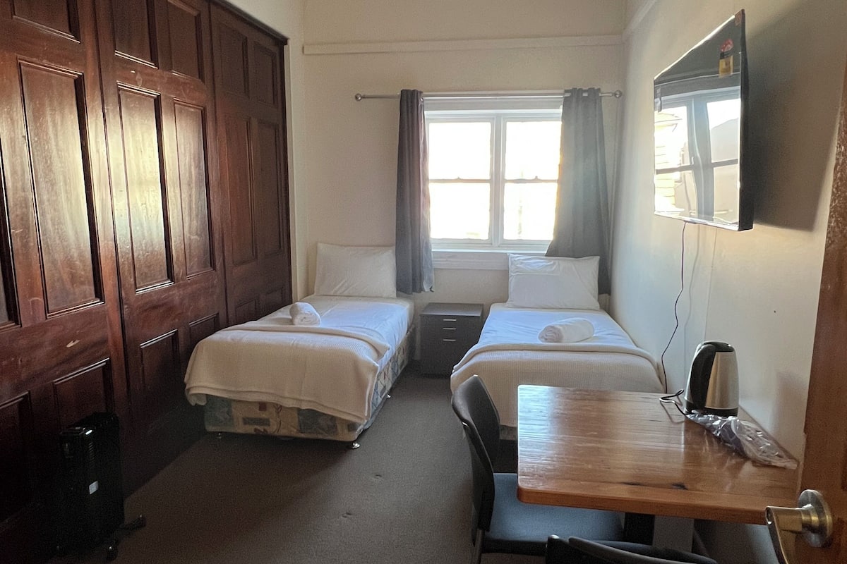 A hotel room with two single beds, a wooden wardrobe, a desk with a chair, a small table, and a TV mounted on the wall.