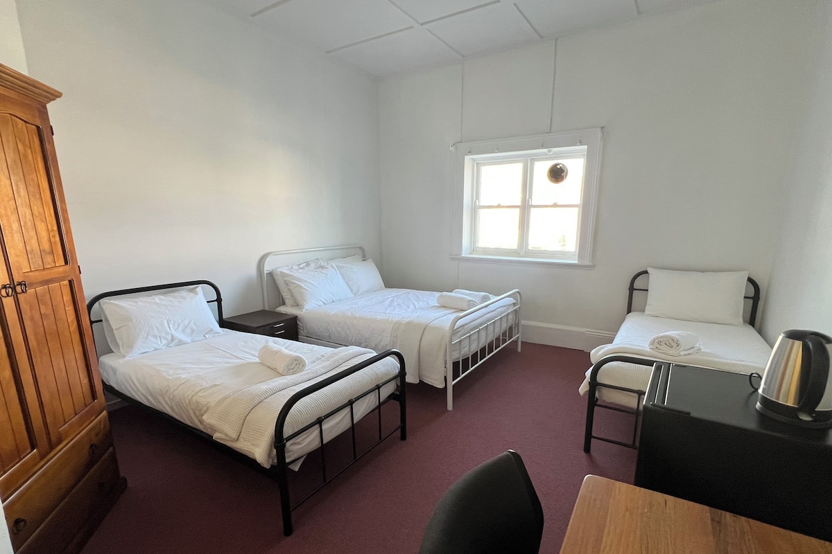 A room with two single beds, a wooden wardrobe, a desk with a kettle, and a window with a view.
