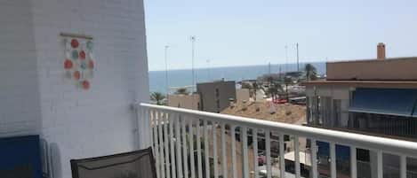 A balcony with a white railing, a table with a candle and a cup, and a view of the sea and buildings in the background.