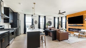 A modern kitchen with a central island, black cabinetry, stainless steel appliances, and a large TV mounted on a wooden wall.