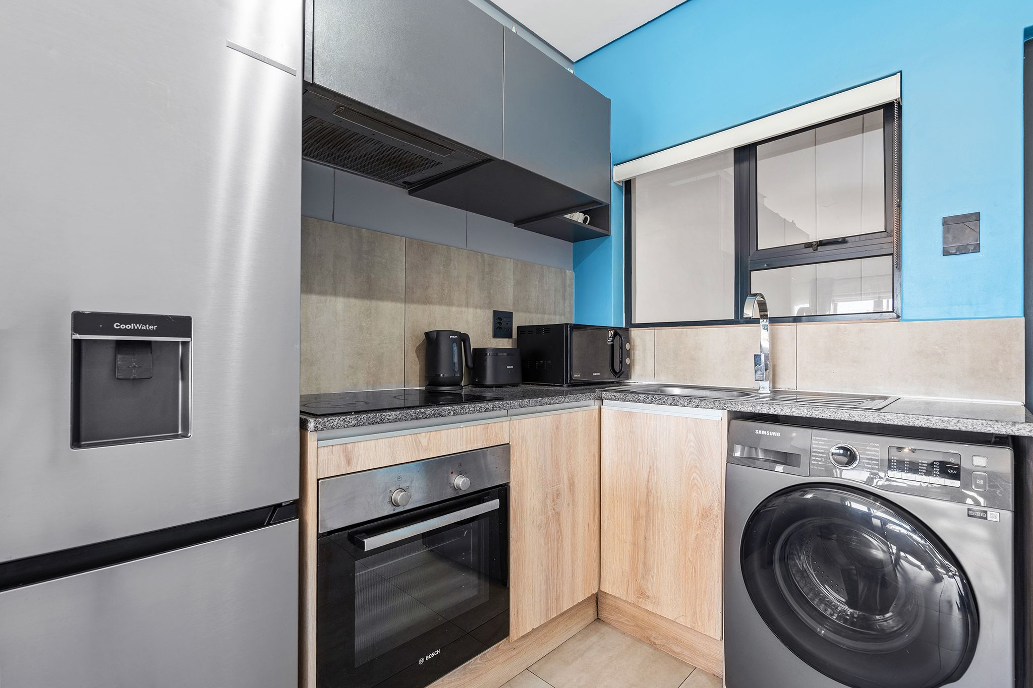 Una cocina compacta con refrigerador de acero inoxidable, horno Bosch, lavadora y cafetera.