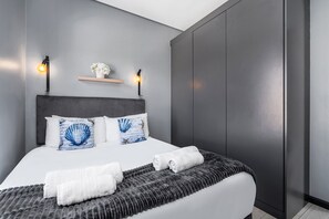 A modern bedroom with a grey headboard, white bedding, a grey textured throw blanket, and two wall-mounted light fixtures. - Blouberg Mews (Cape Town)