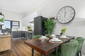 A modern kitchen with a large wall clock, a dining area with a wooden table, and green upholstered chairs. - Atlantic Port 10 (Cape Town)