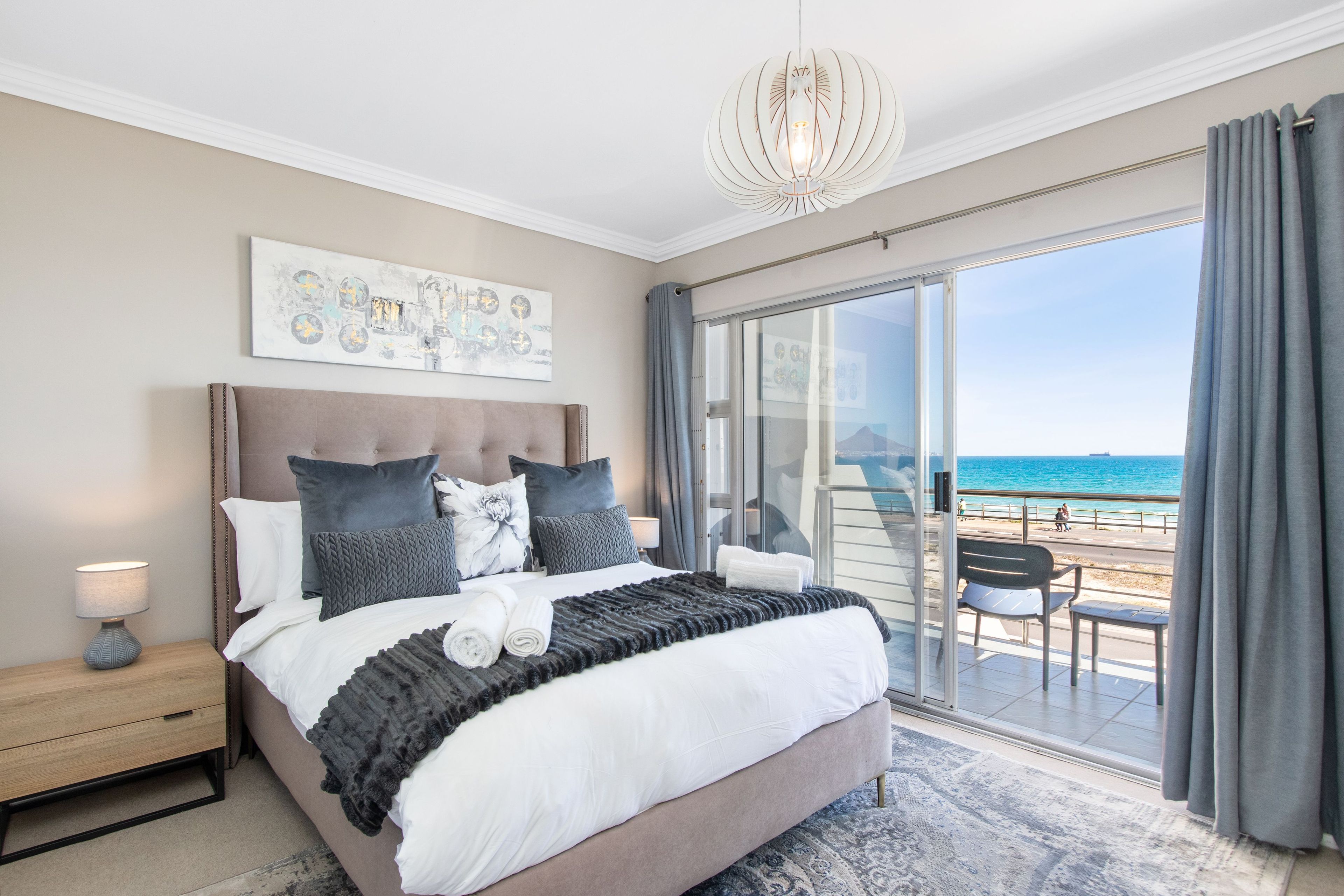 Een slaapkamer met een groot bed, een nachtkastje, een plafondlamp en uitzicht op het strand door een schuifdeur.