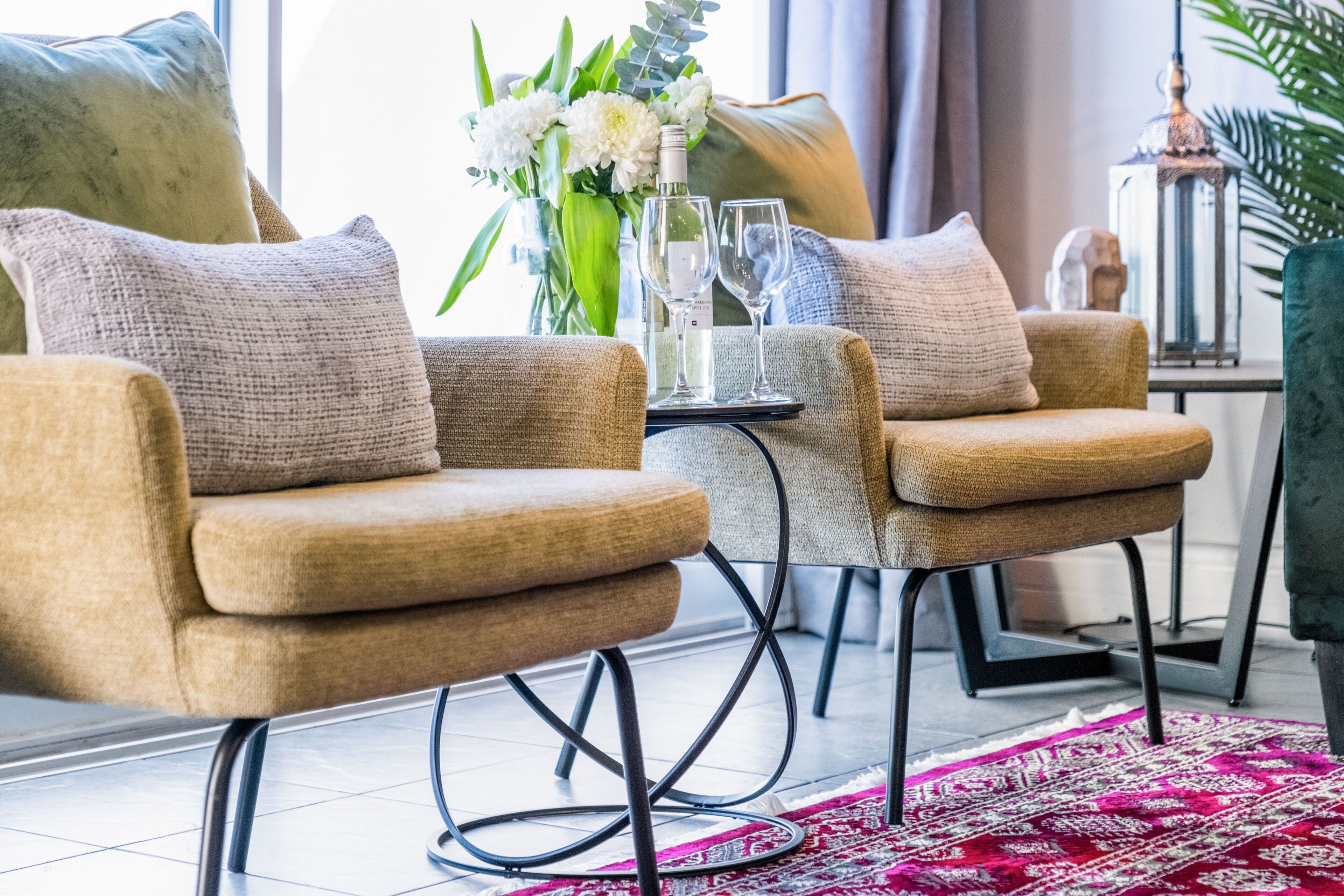 A modern living room with two beige armchairs, a small side table with a vase of flowers, and a patterned rug.