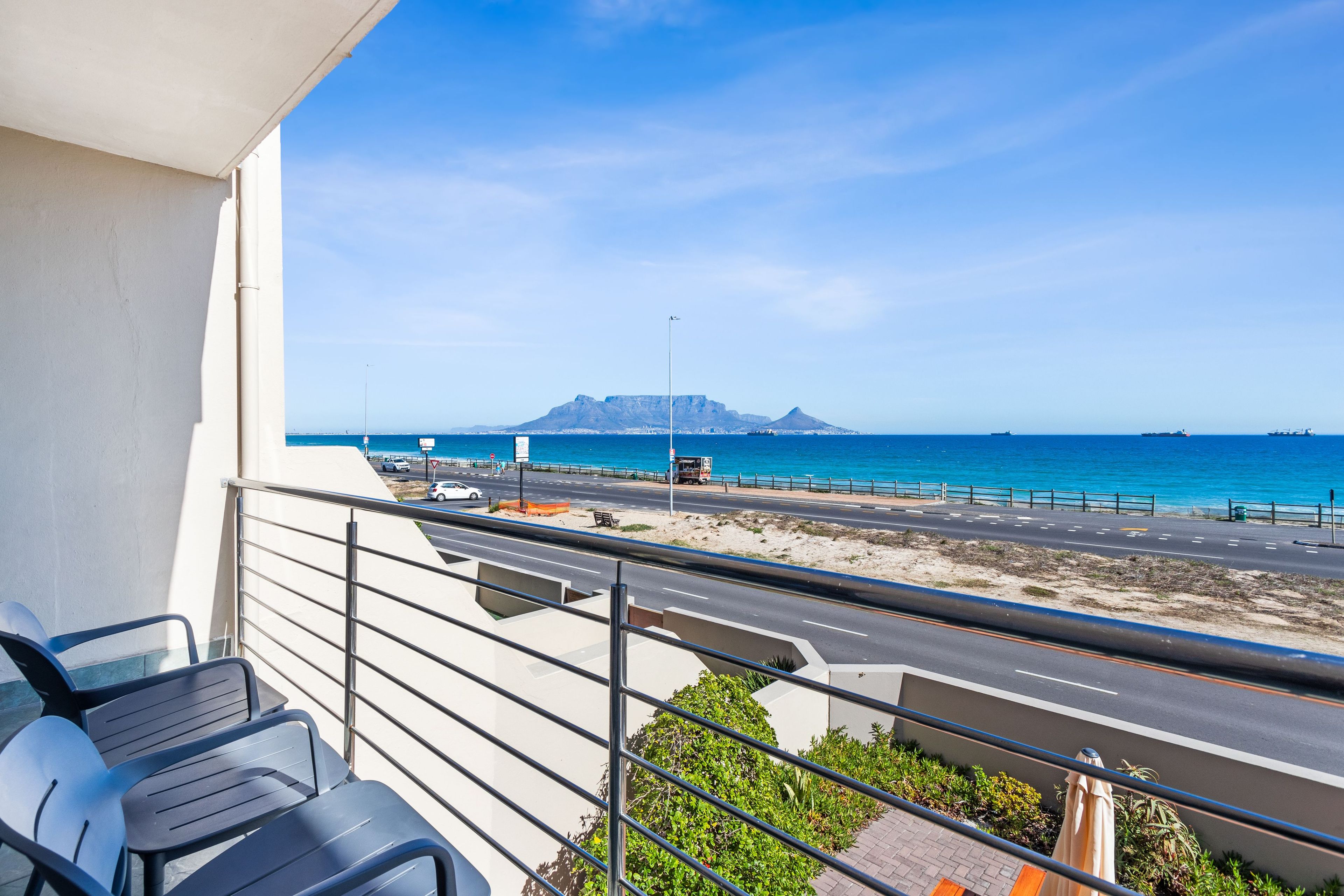 A balcony with chairs overlooking a beach and road, with a mountain in the distance.