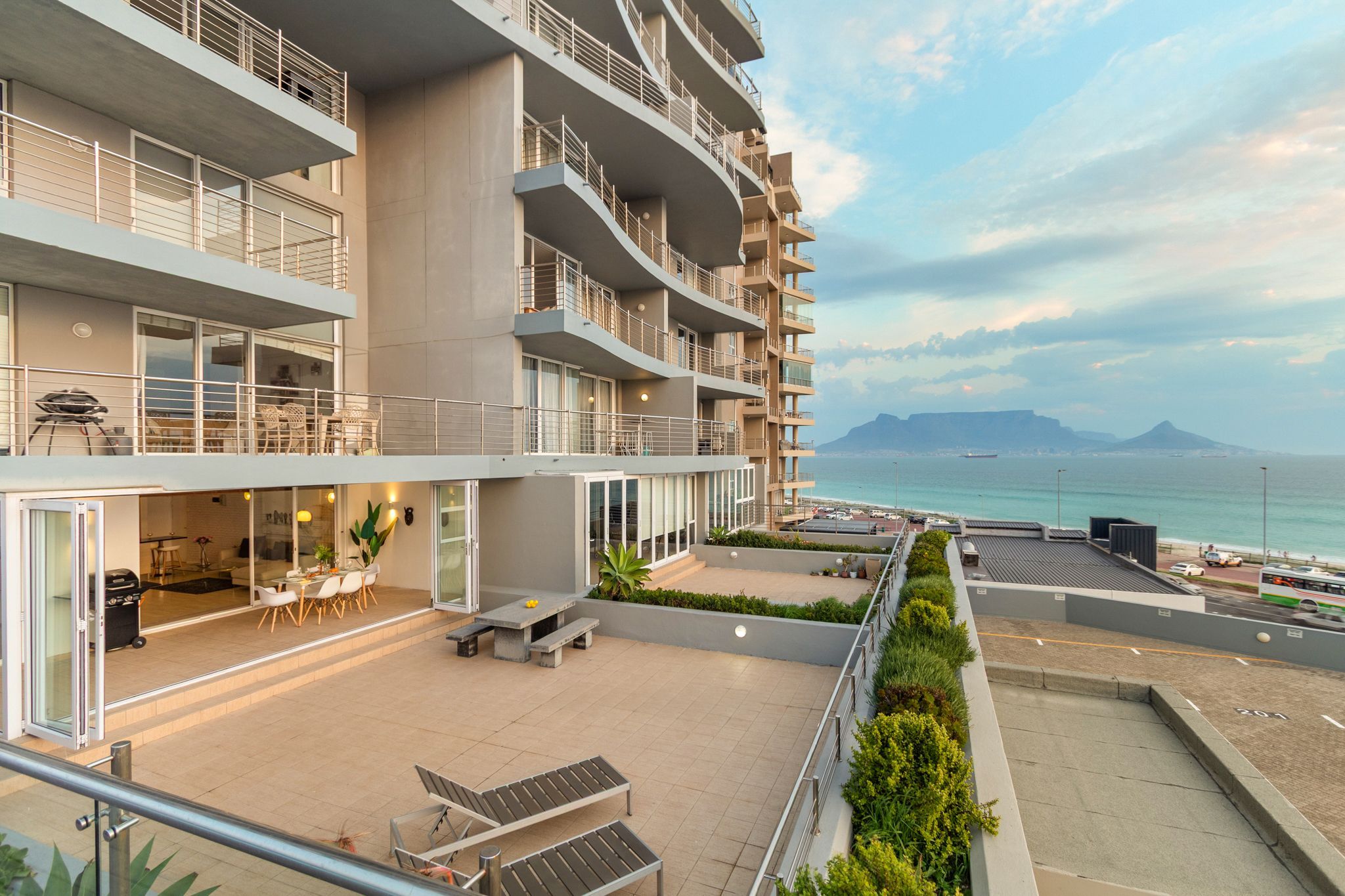 A modern multi-story hotel with balconies, outdoor seating, and a view of the sea.