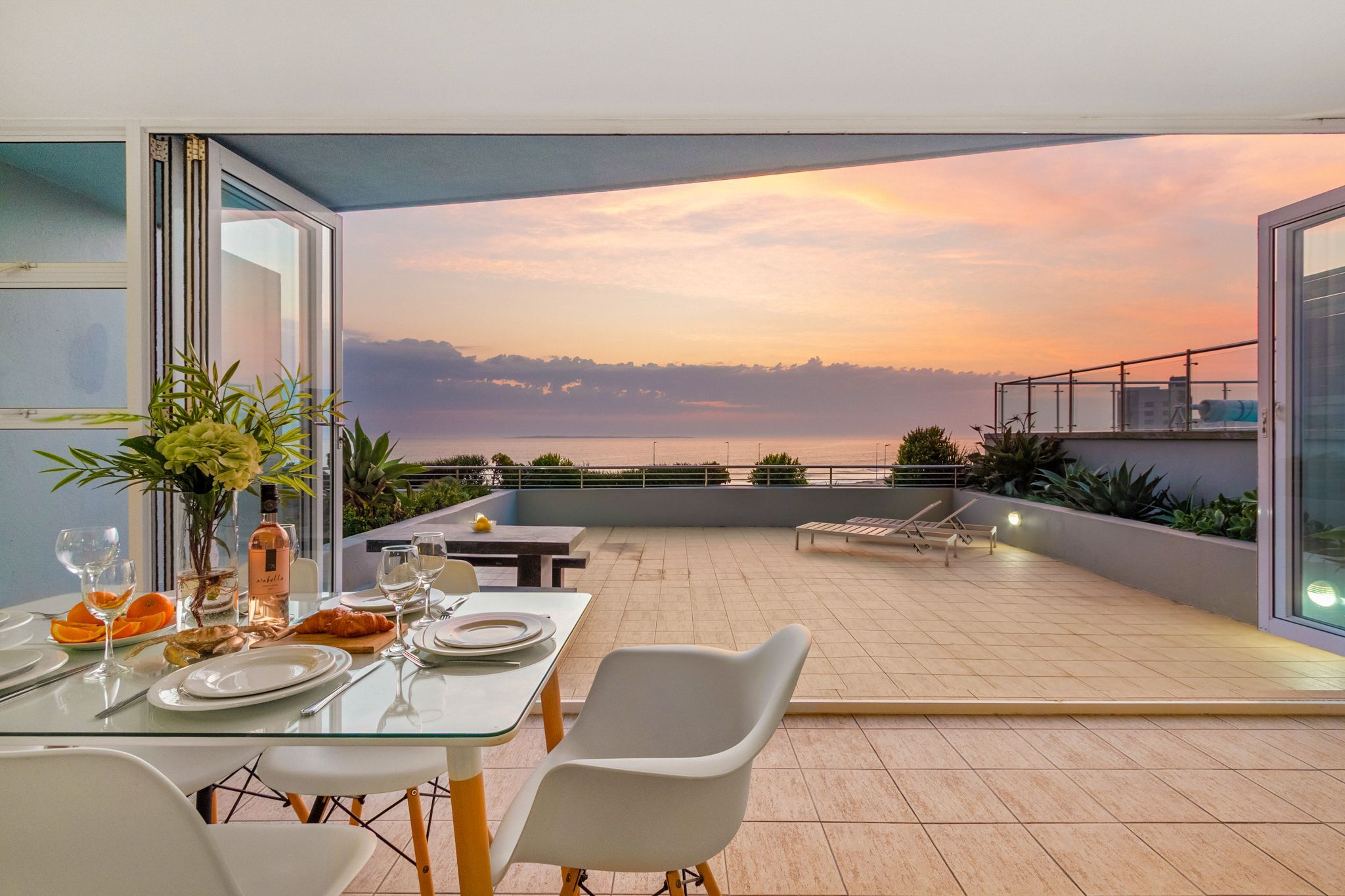 A dining area with a table set for two, overlooking a scenic view of the sea at sunset.