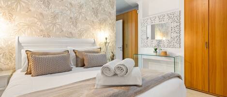 A bedroom with a large bed, a glass bedside table, a wooden wardrobe, and a decorative mirror.