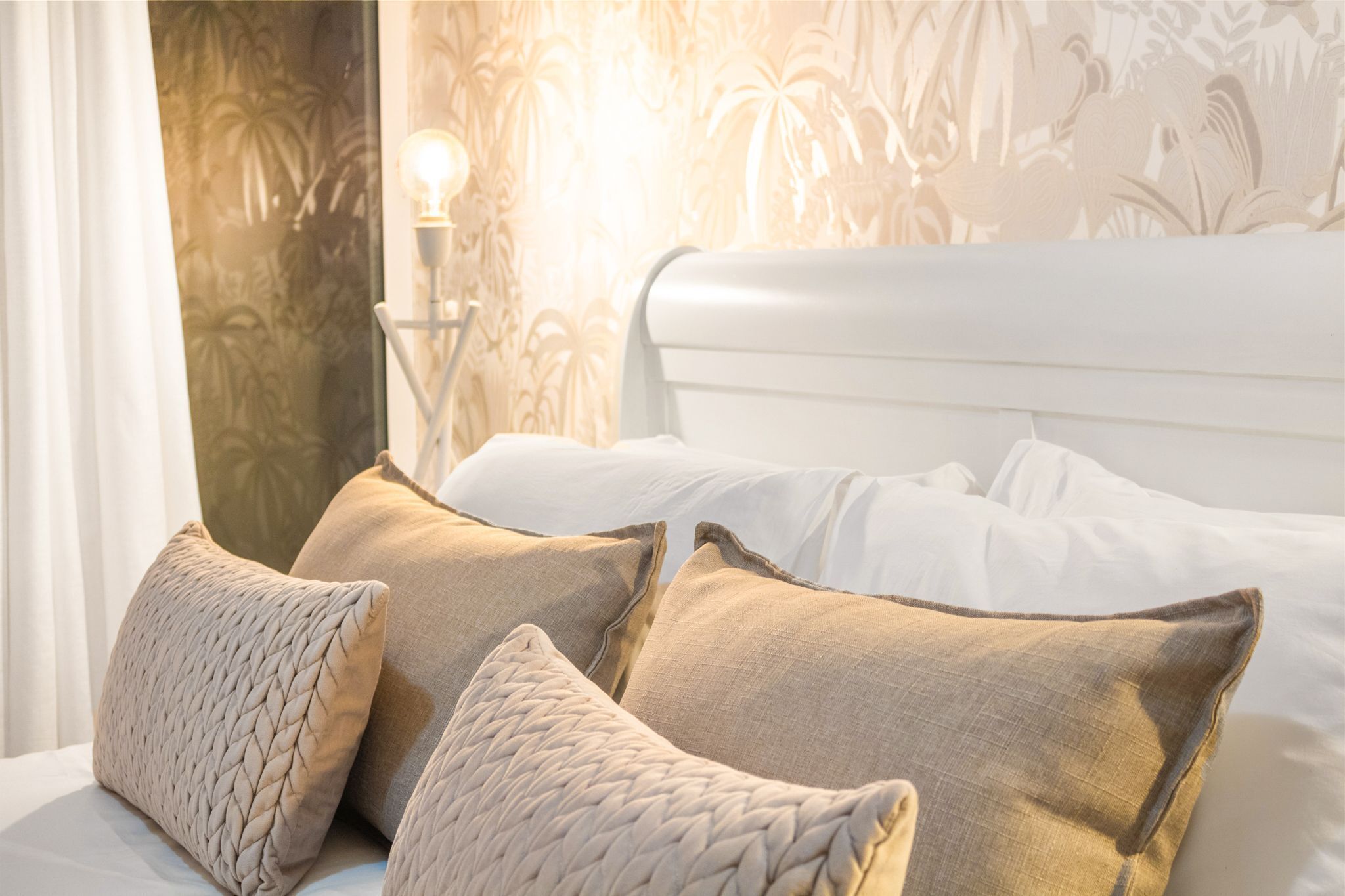 A neatly made bed with white linens and beige pillows, a wall with a floral pattern, and a wall-mounted light fixture.