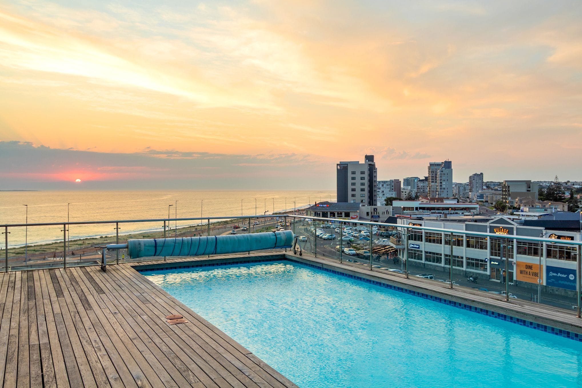 Une piscine dotée d’une terrasse en bois, offrant une vue sur le paysage urbain et l’océan au coucher du soleil.