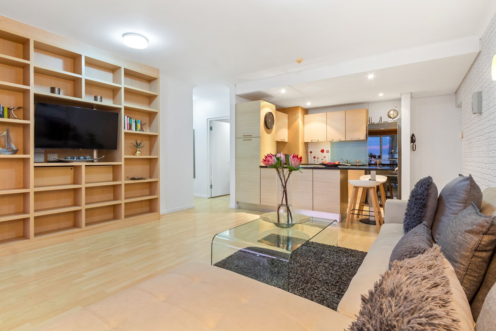 A modern living room with a glass coffee table, a flat-screen TV, wooden shelving, and a kitchen in the background.