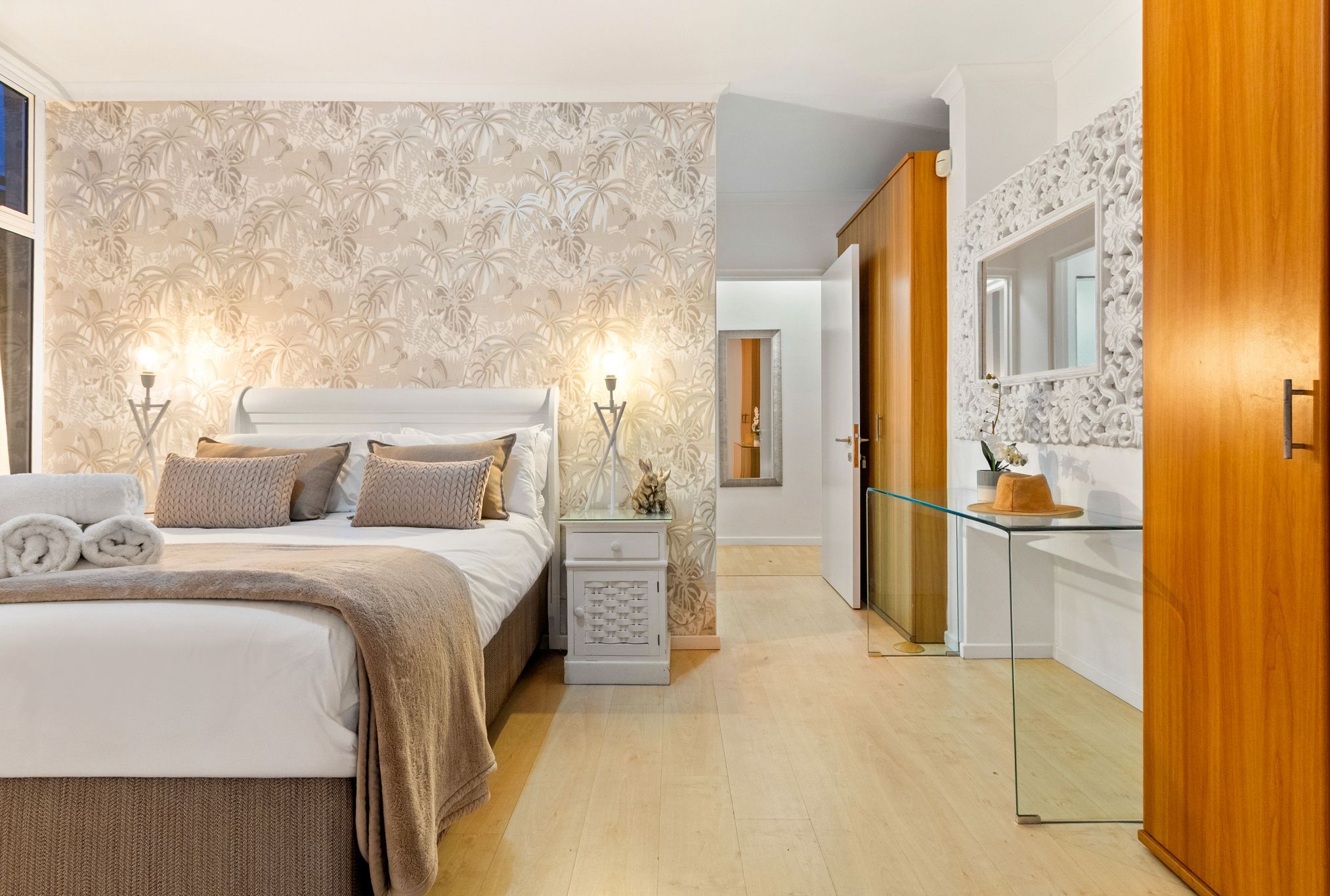A bedroom with a bed, bedside tables, and a nightstand. There is a mirror above the nightstand and a door leading to another room.