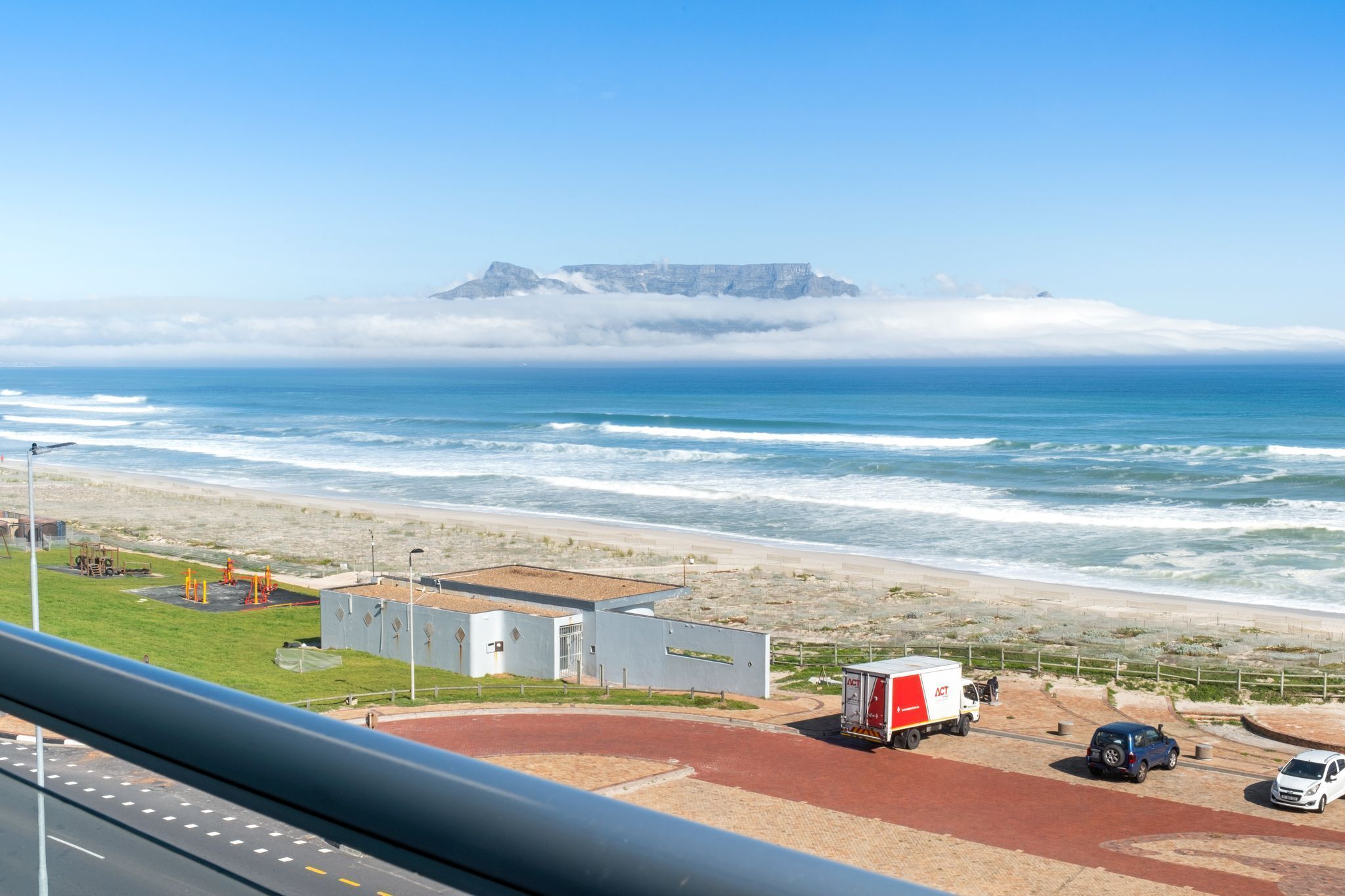 A coastal view with a beach, a truck, and a car parked near a building with a flat roof.