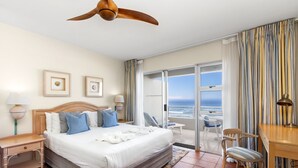 Quarto de hotel com cama, escrivaninha, cadeira, ventoinha no teto e vista para o oceano.