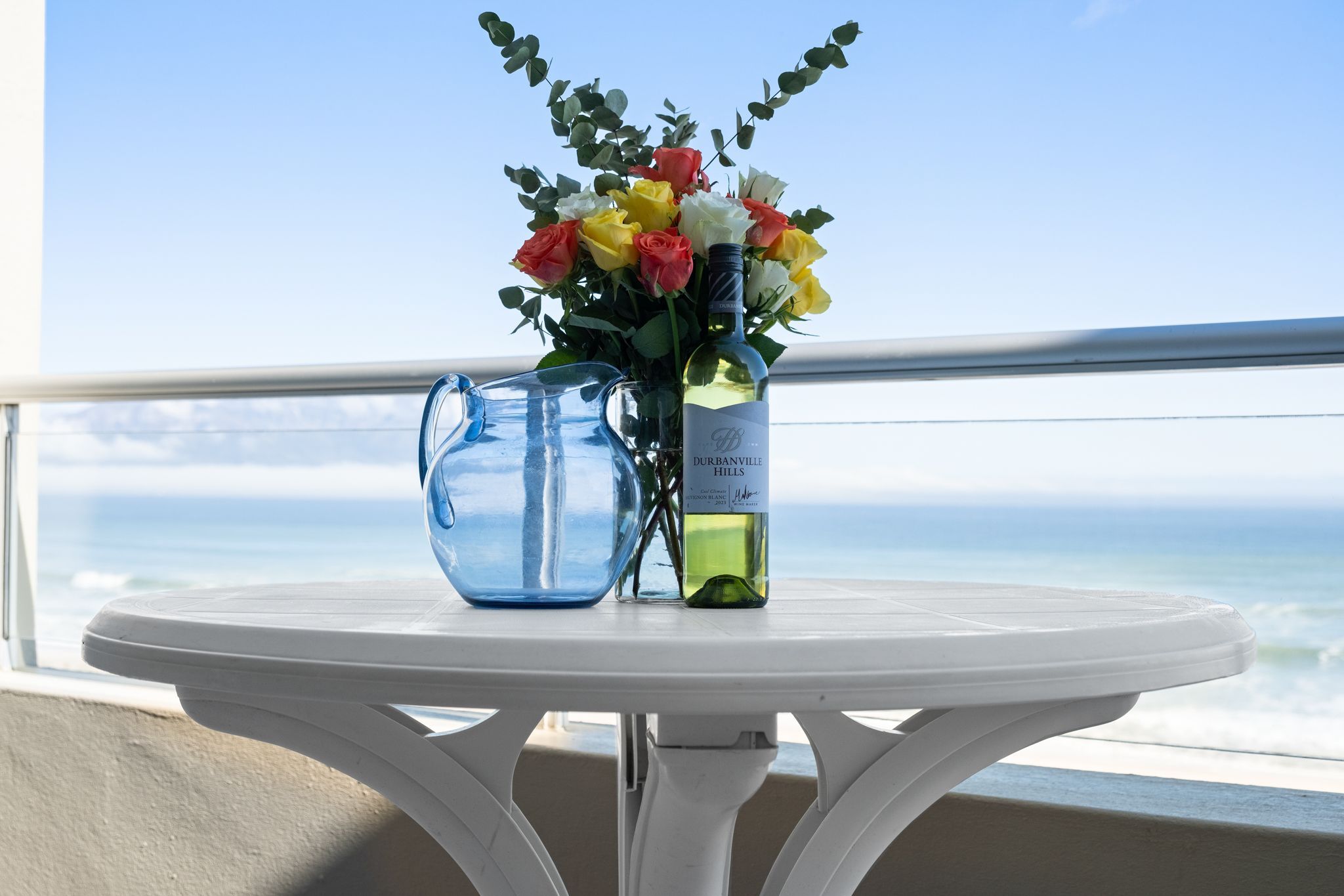 A table with a vase of flowers and a bottle of wine on a balcony overlooking the ocean.
