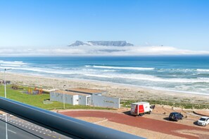 A coastal view with a beach, a truck, and a car parked near a building with a flat roof.