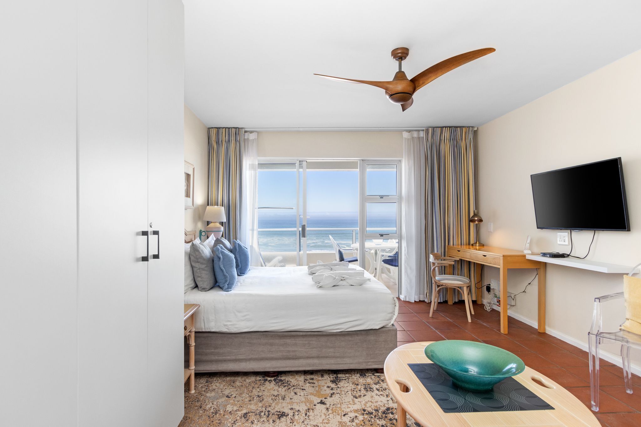 A hotel room with a bed, a desk, a TV, and a view of the ocean.