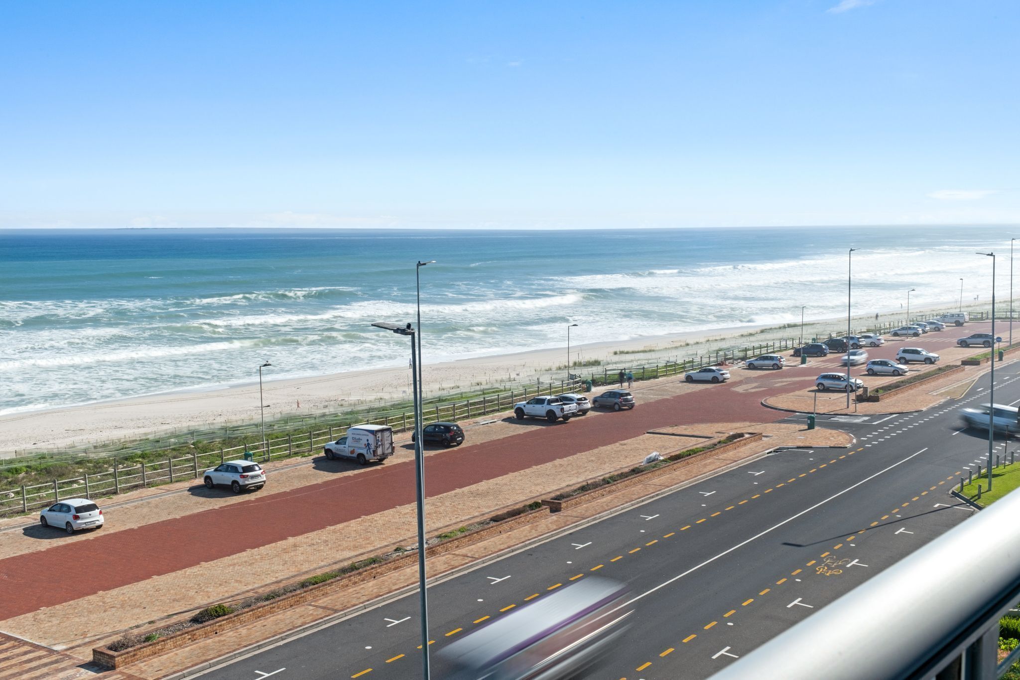A coastal road with a red bike lane, a beach, and parked cars.