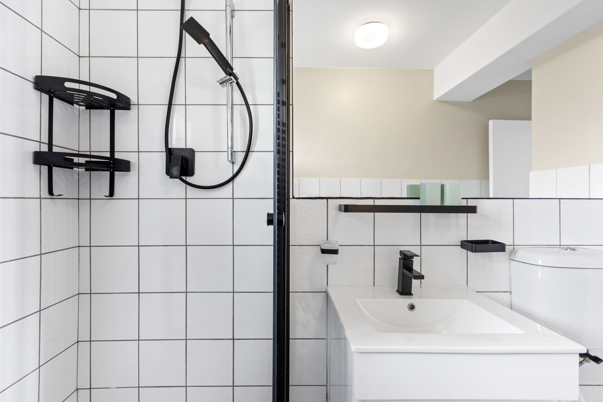 A modern bathroom with white tiled walls, a black showerhead, a white sink, and a mirror above the sink.