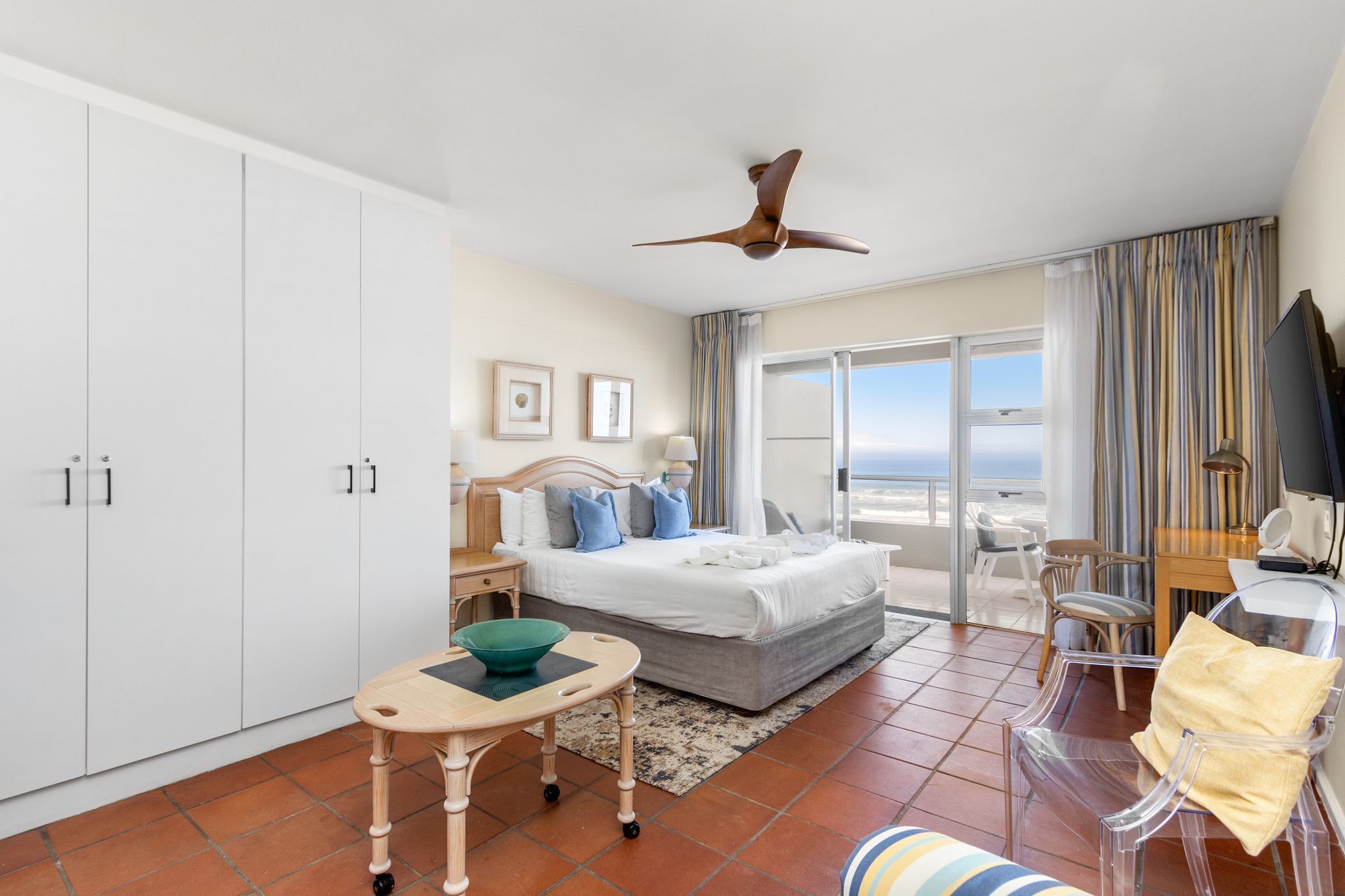 A hotel room with a bed, a desk, a chair, a ceiling fan, and a view of the ocean.
