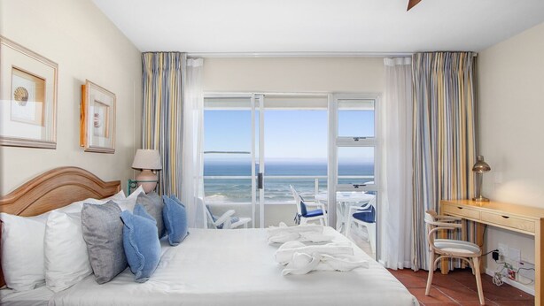 Quarto de hotel com cama grande, escrivaninha, cadeira e vista para o oceano.
