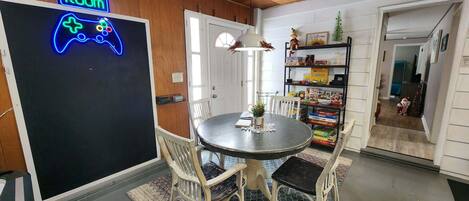 A room with a neon sign reading "GAME ROOM," a round table with chairs, a small plant, and a bookshelf filled with board games.