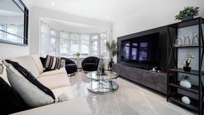 A modern living room with a sofa, armchairs, a glass coffee table, a TV, and a bookshelf.