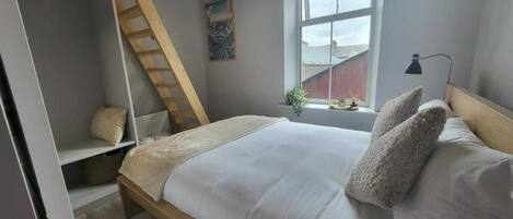 A bedroom with a loft bed, a window with a view, a ladder, and a bedside lamp.