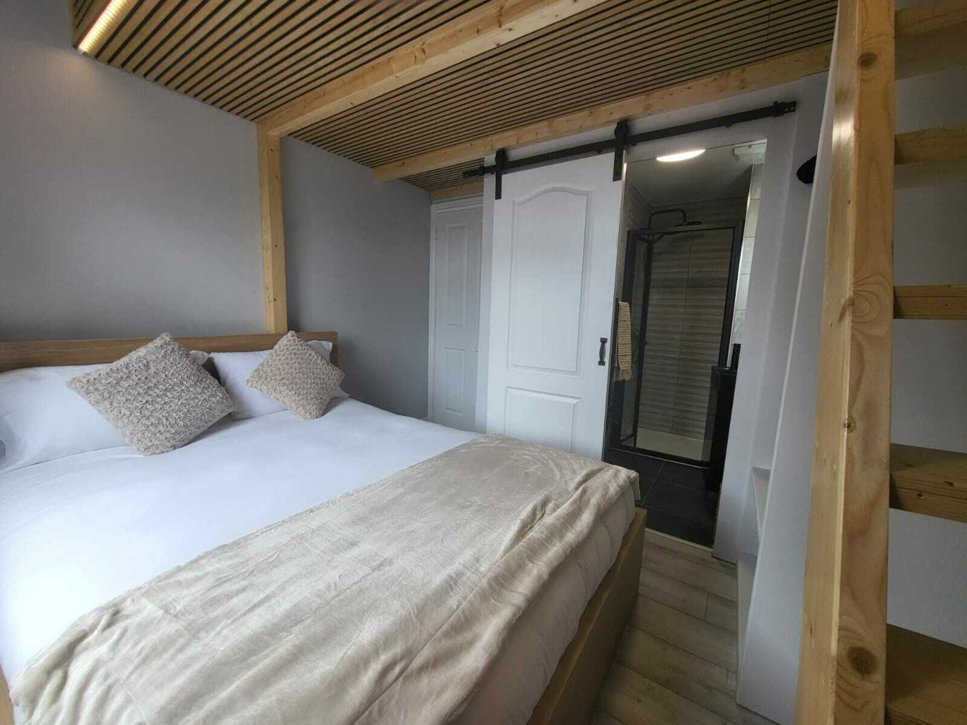 A bedroom with a bed, two pillows, a wooden ceiling, a white door, and a shower area.