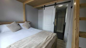 A bedroom with a bed, two pillows, a wooden ceiling, a white door, and a shower area.