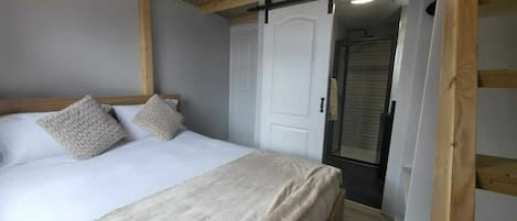 A bedroom with a bed, two pillows, a wooden ceiling, a white door, and a shower area.