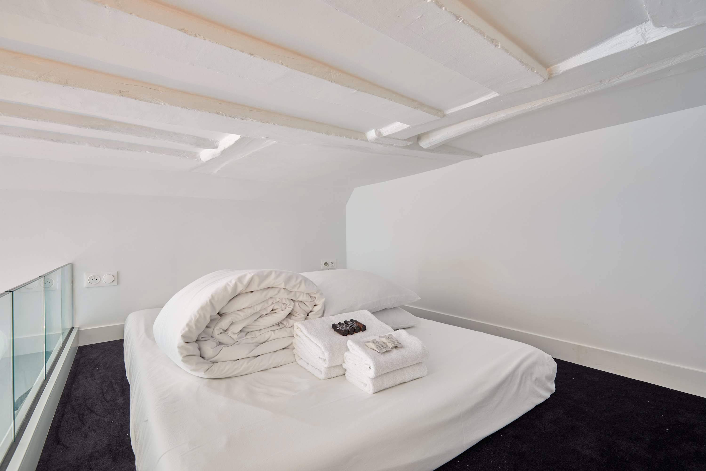 A bedroom with a bed, white bedding, a glass railing, and a black carpet.
