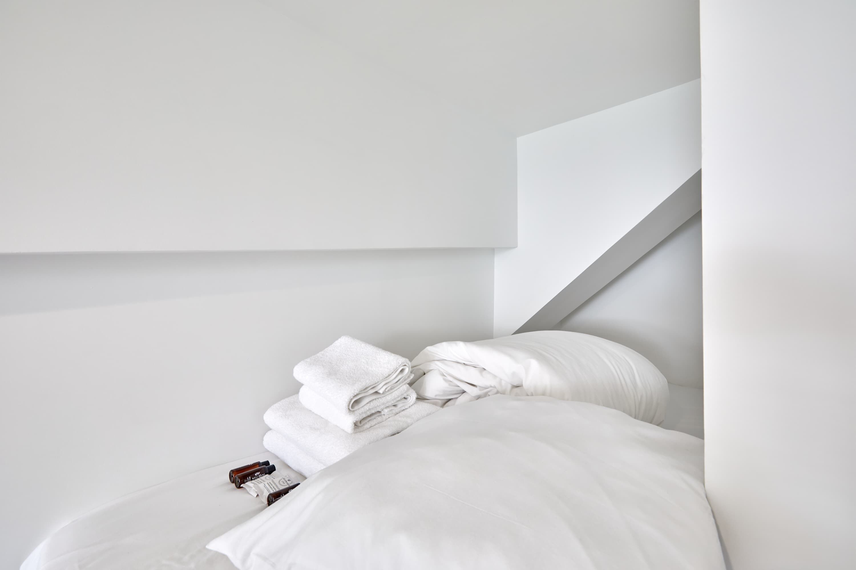 Un lit bien fait, avec des draps blancs et une serviette pliée, un oreiller blanc et une petite bouteille posée à côté.