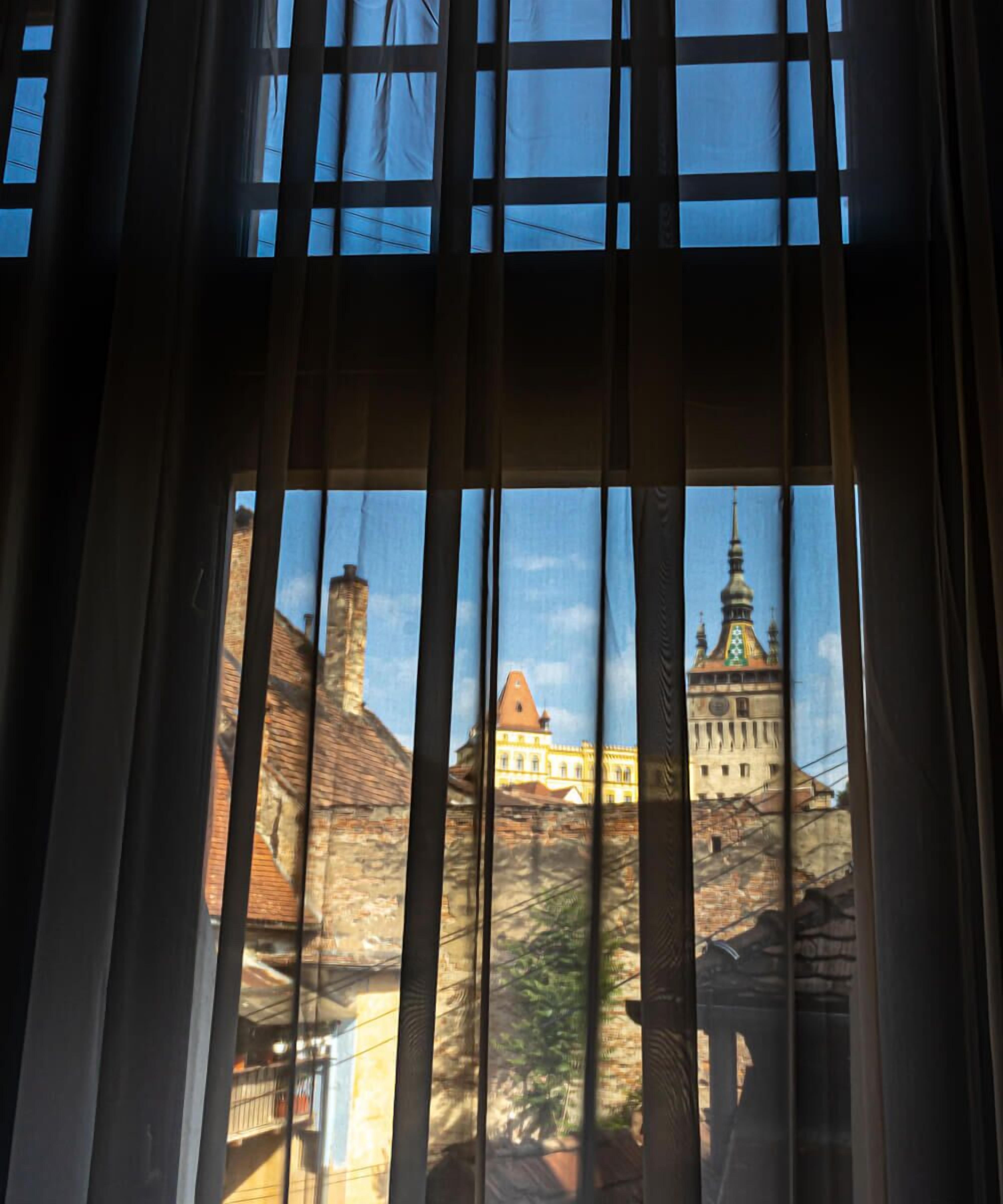 A view through a window with vertical blinds, featuring a building with a prominent tower and a smaller building with a red roof in the background.