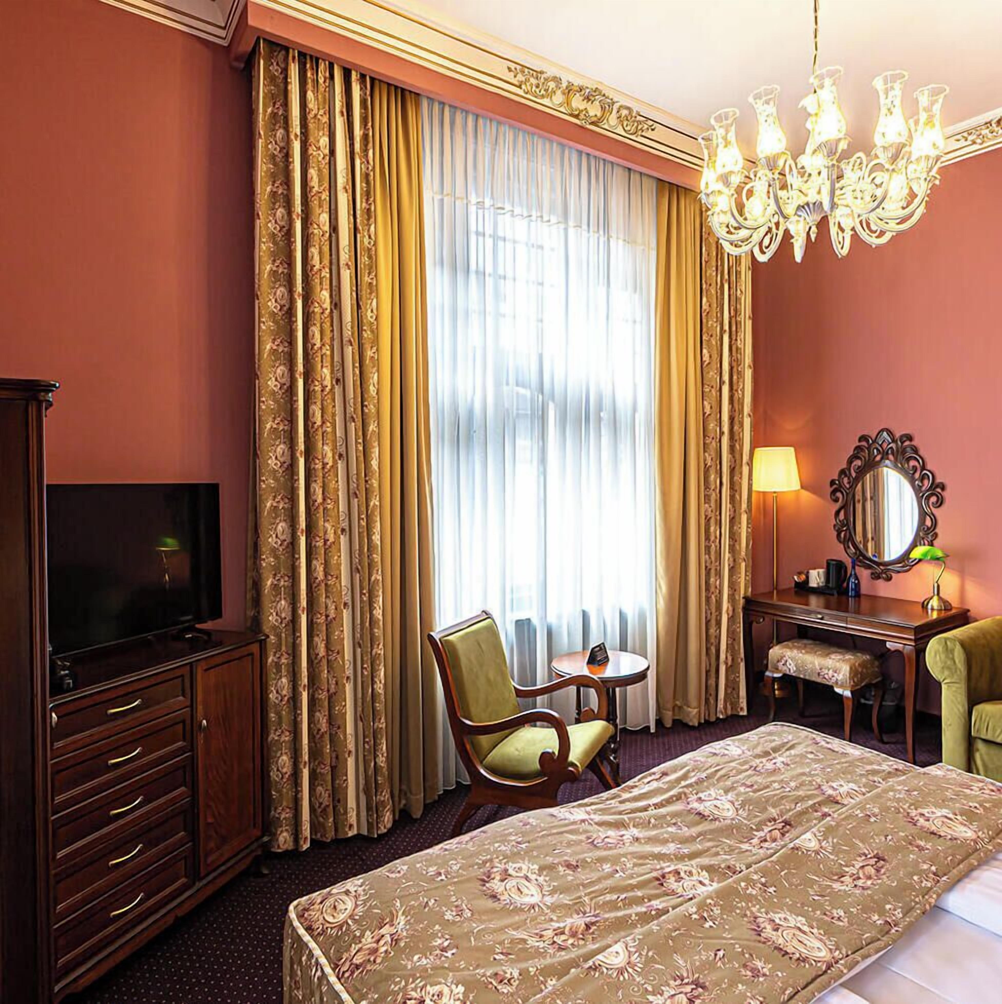 A hotel room with a bed, a television, a desk with a lamp, a chair, and a large window with curtains.