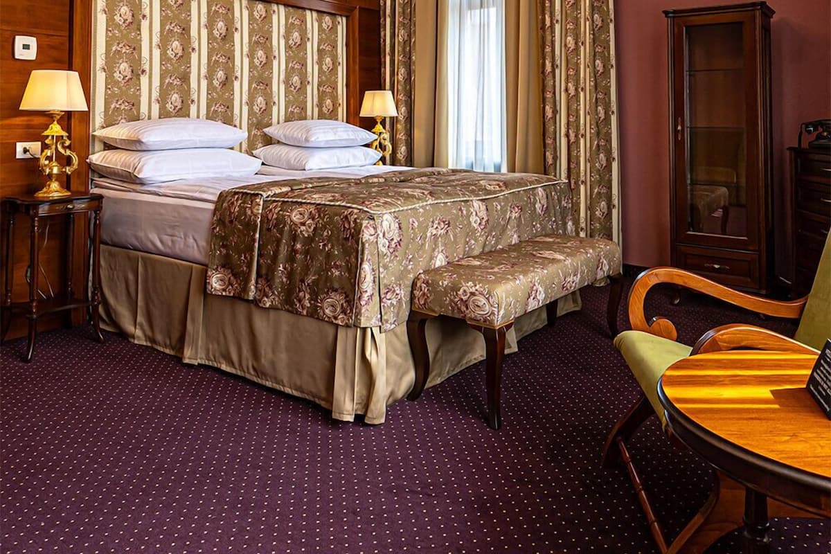 A hotel room with a large bed, two bedside tables, a wooden wardrobe, a chandelier, and a dining table with chairs.
