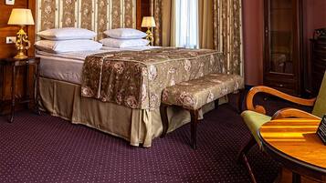 A hotel room with a large bed, two bedside tables, a wooden wardrobe, a chandelier, and a dining table with chairs.