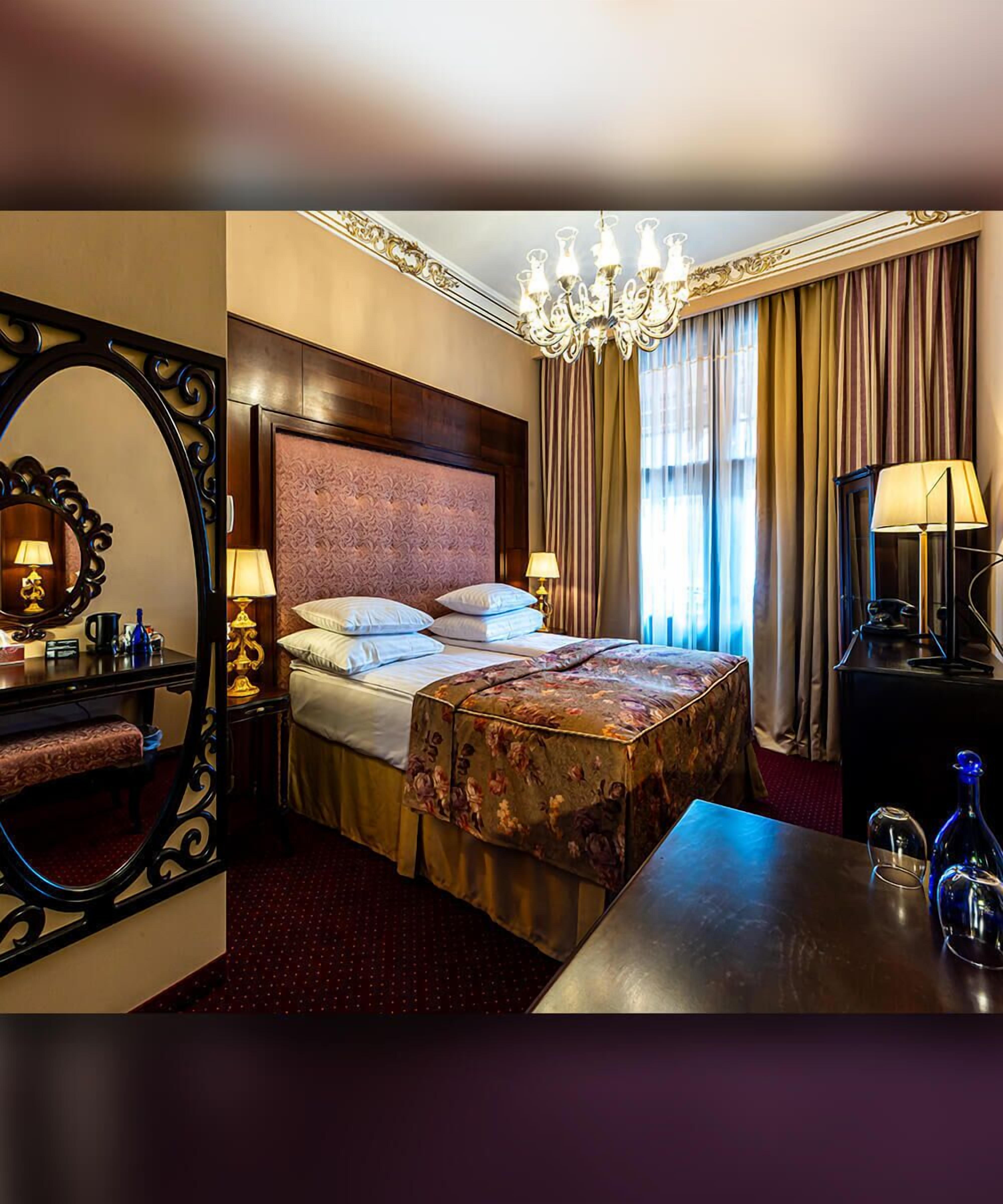A hotel room with a large bed, a wooden headboard, a chandelier, a desk with a lamp, and a mirror.