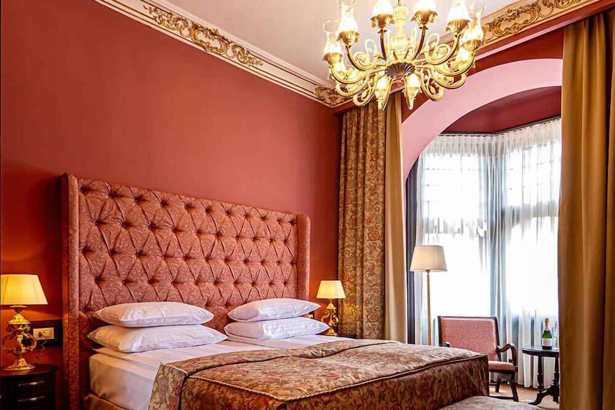 A hotel room with a tufted headboard bed, a bench, two bedside tables with lamps, a chandelier, and a view of the city through large windows.