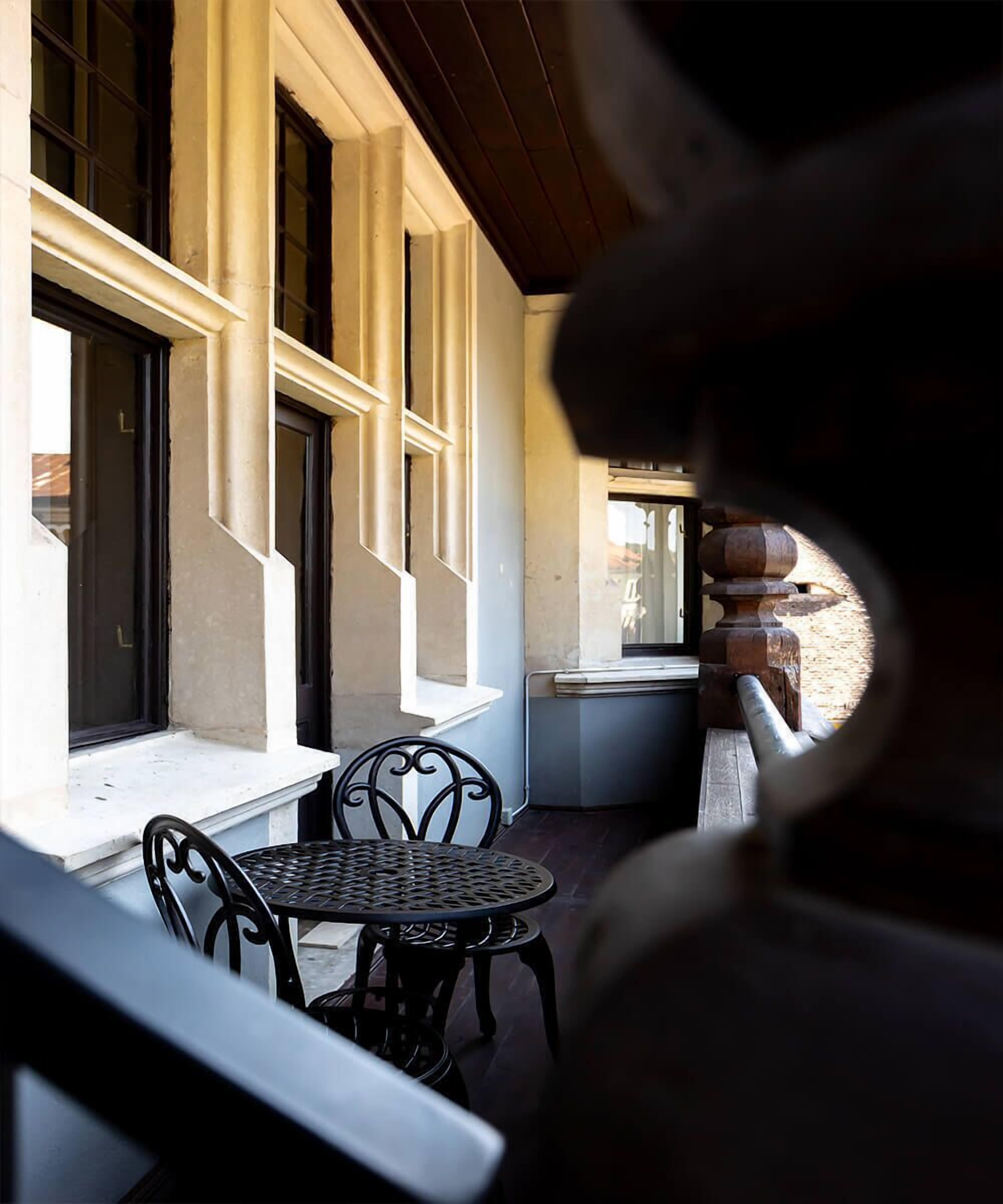A covered outdoor seating area with a table and chairs, a window with a view of the street, and a stone pillar.