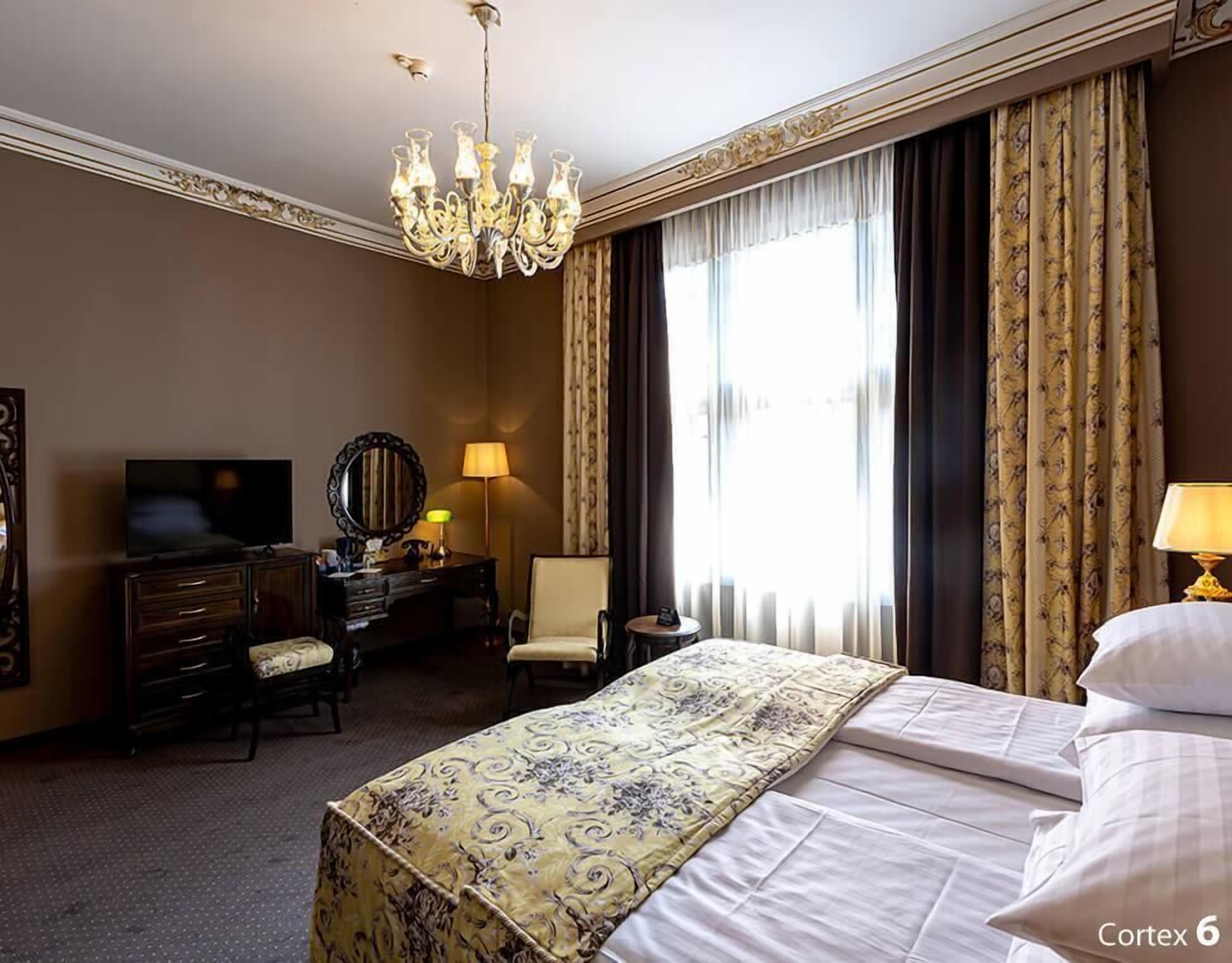 A hotel room with a large bed, a television, a desk with a chair, a dresser, and a chandelier.