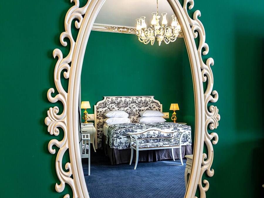 Un dormitorio con un espejo grande y ornamentado, una cama con ropa de cama floral, dos mesitas de noche con lámparas y un candelabro.