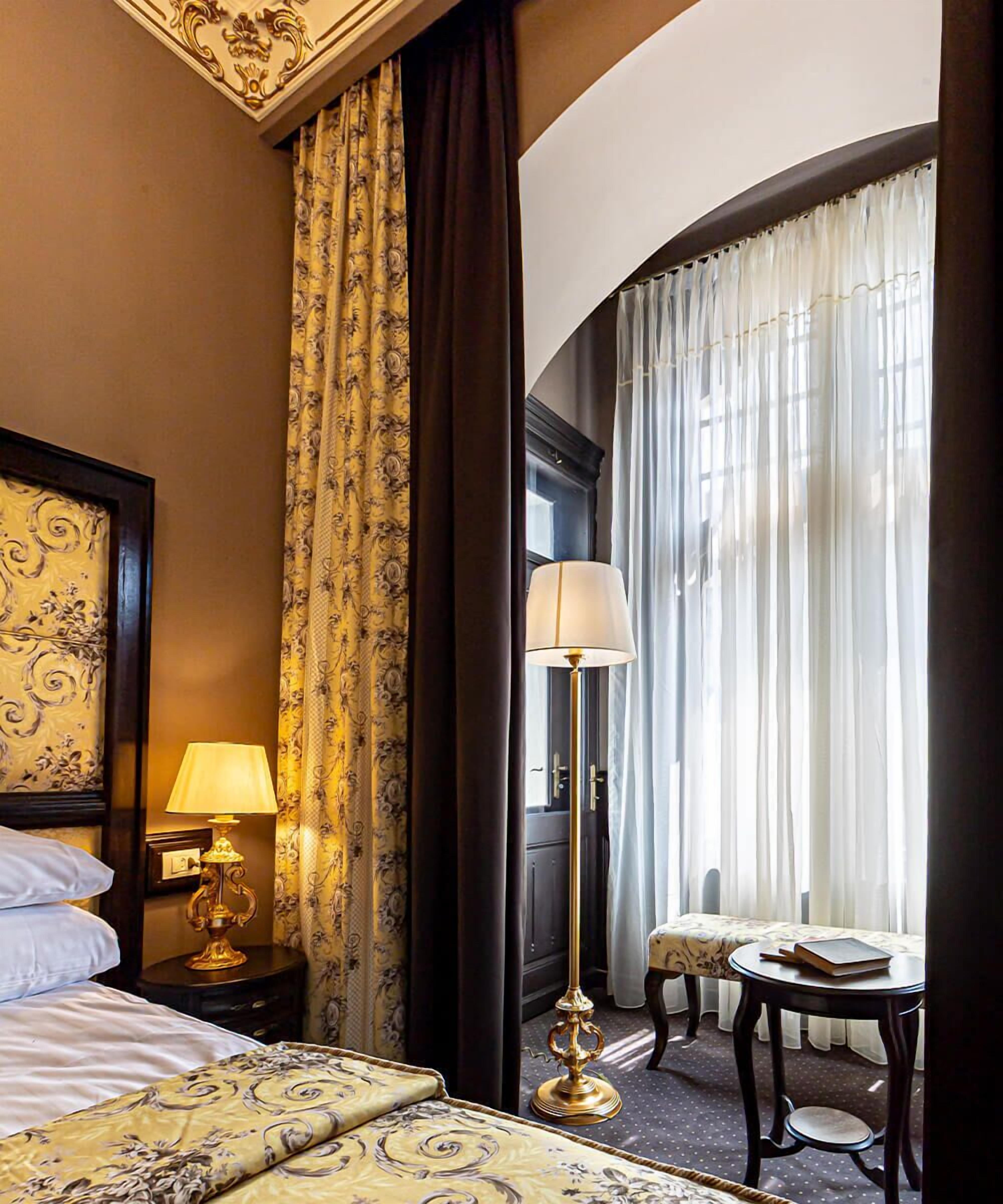 A hotel room with a bed, a nightstand with a lamp, a sitting area with a table and chair, and a large window with curtains.
