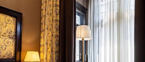 A hotel room with a bed, a nightstand with a lamp, a sitting area with a table and chair, and a large window with curtains.