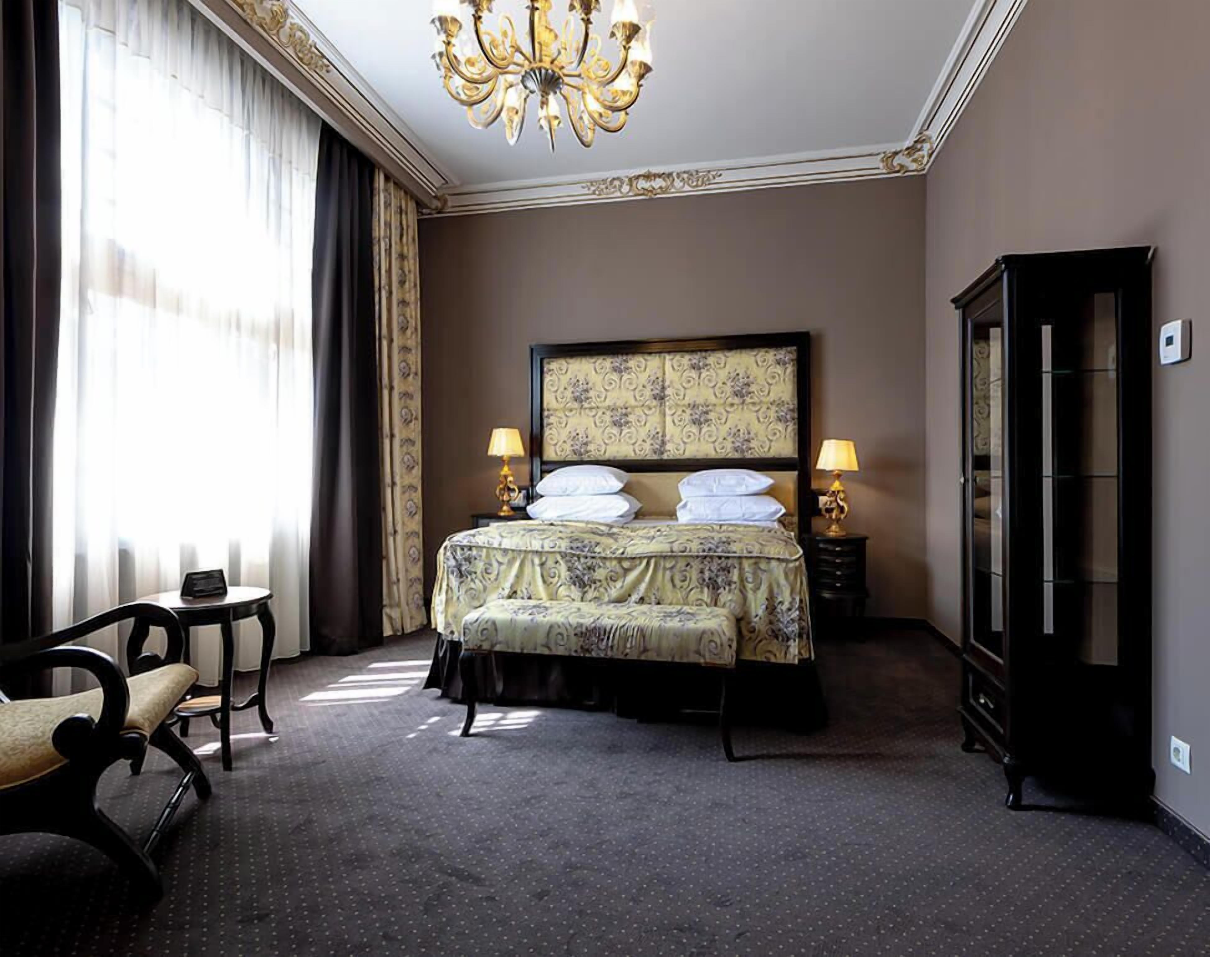 Habitación de hotel con una cama grande, dos mesitas de noche, una araña, un armario y un espacio de descanso con una silla y una pequeña mesa.