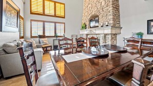 Un comedor amplio con una gran mesa de madera, chimenea de piedra y varios ventanales con persianas.
