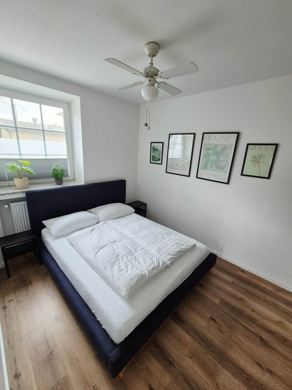 A bedroom with a bed, a ceiling fan, a window with blinds, a radiator, and framed artwork on the wall. - 2-room apartment 1 - Apartments am Schloss (Oberschleißheim)