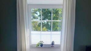 A dining area with a glass table and two chairs, a window with white curtains, and a view of greenery outside. - 2-room apartment 1 - Apartments am Schloss (Oberschleißheim)