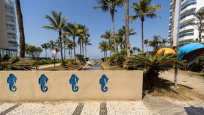 A coastal walkway with palm trees, a beach, and a building with balconies. - Apto em Riviera de São Lourenço a 300m da Praia (Bertioga)